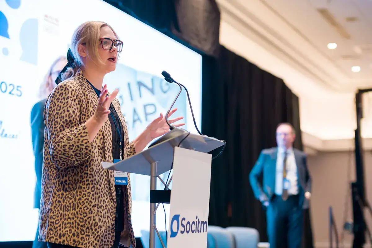 A woman in a leopard-print jacket speaks at a lectern with a Socitm sign, gesturing with her hands. A man in a suit stands blurred in the background on a brightly lit stage.
