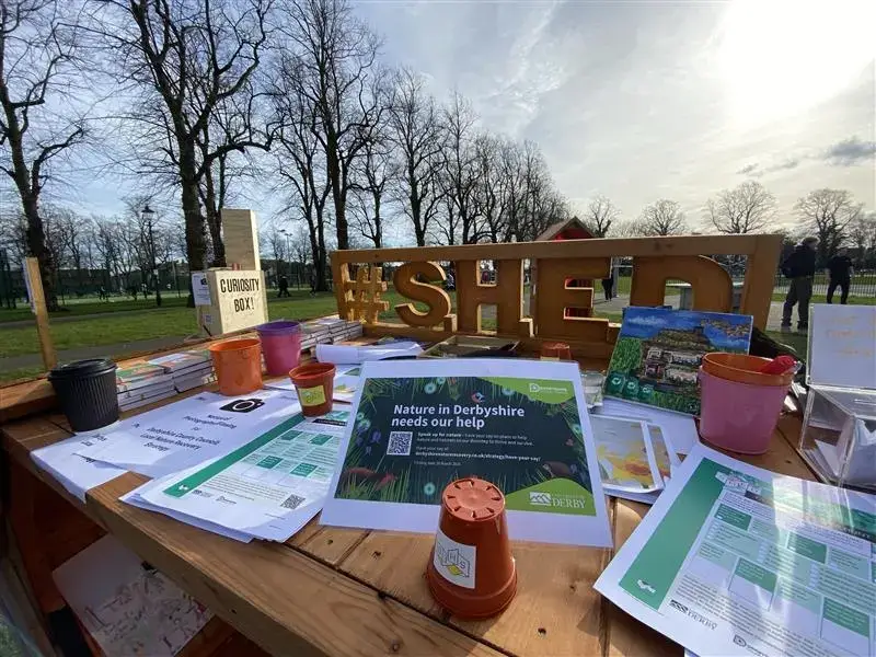 A wooden outdoor table displays gardening leaflets, small plant pots, and paperwork, with a large #SHED sign. Leafless trees and a park with green grass are visible in the background under a partly cloudy sky.