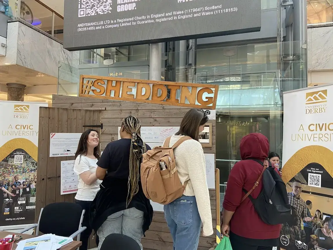 Four people stand at an indoor stall labelled #SHEDDING with posters and information. Two University of Derby banners are visible, and a large digital screen is in the background.