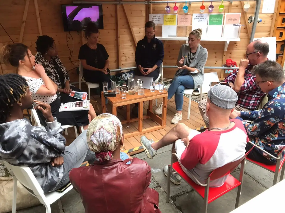 A group of people sit in a circle inside a wooden room, having a discussion. Some hold papers, whilst others listen. Drinks are on a small table in the centre, and colourful labels hang on the wall behind them.