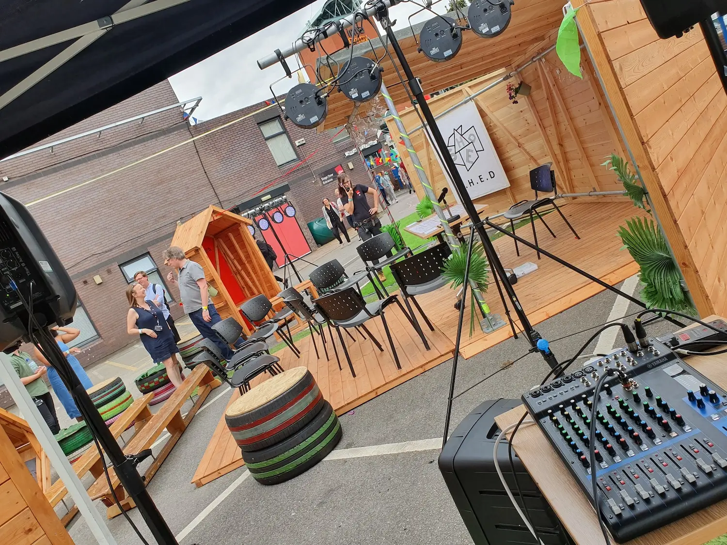 A small outdoor stage set-up includes chairs, microphones, and audio equipment, with wooden structures and colourful decorations. People stand and chat in the background near a brick building.