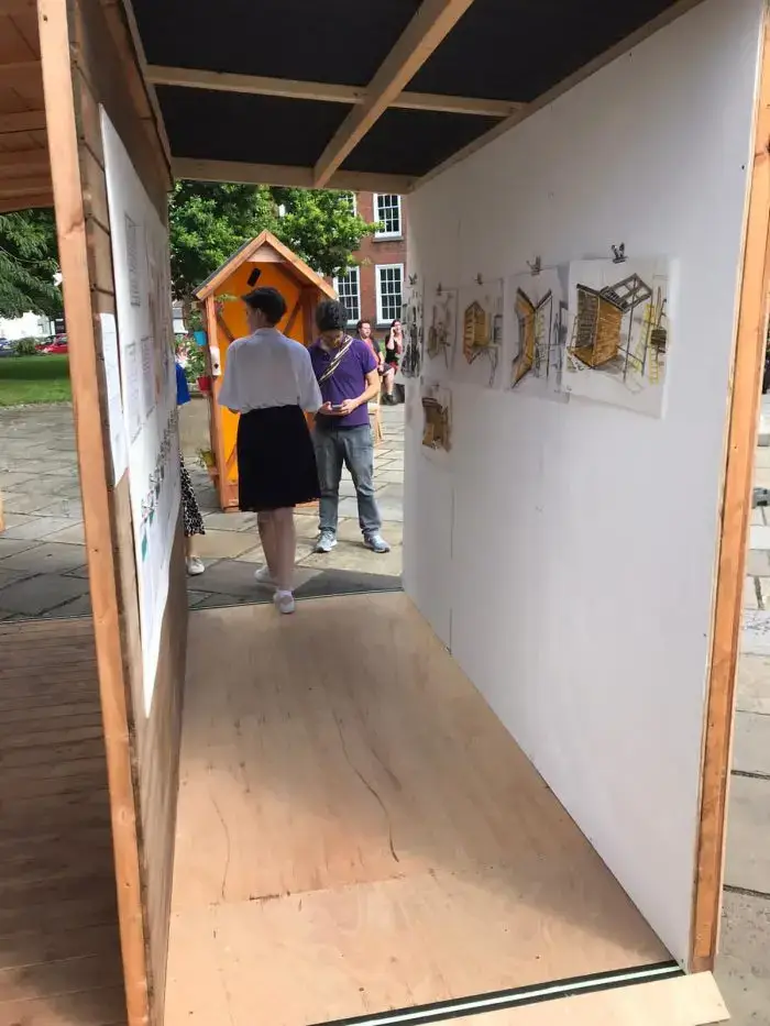 A woman in a white shirt and black skirt walks through a wooden structure displaying architectural drawings; a man stands outside, and people and trees are visible in the background.