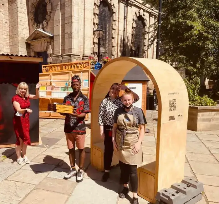 Four people pose outdoors beside a wooden archway structure with QR codes, in front of an old stone building. One person holds a wooden crate, and another wears an apron. It is sunny, and they are smiling at the camera.