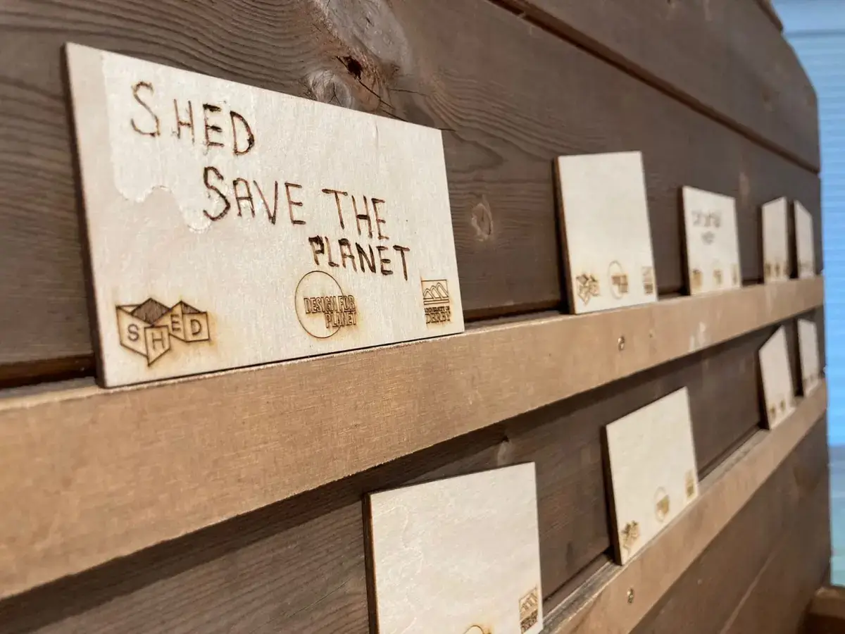 Several wooden plaques displayed on a wall, with one in focus engraved with the words SHED SAVE THE PLANET and small logos underneath. The background shows more similar plaques out of focus.