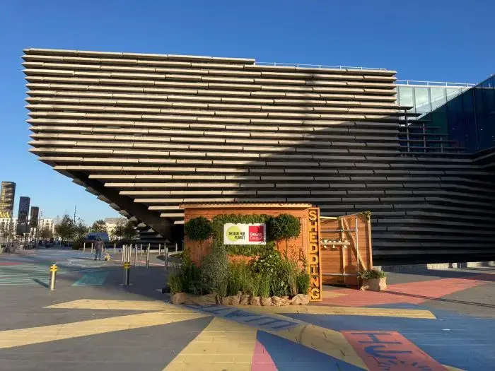 A small wooden shed with plants, labelled SHEDDING, stands in front of a modern, angular building with layered stone-like horizontal slabs under a clear blue sky. Colourful patterns decorate the ground.
