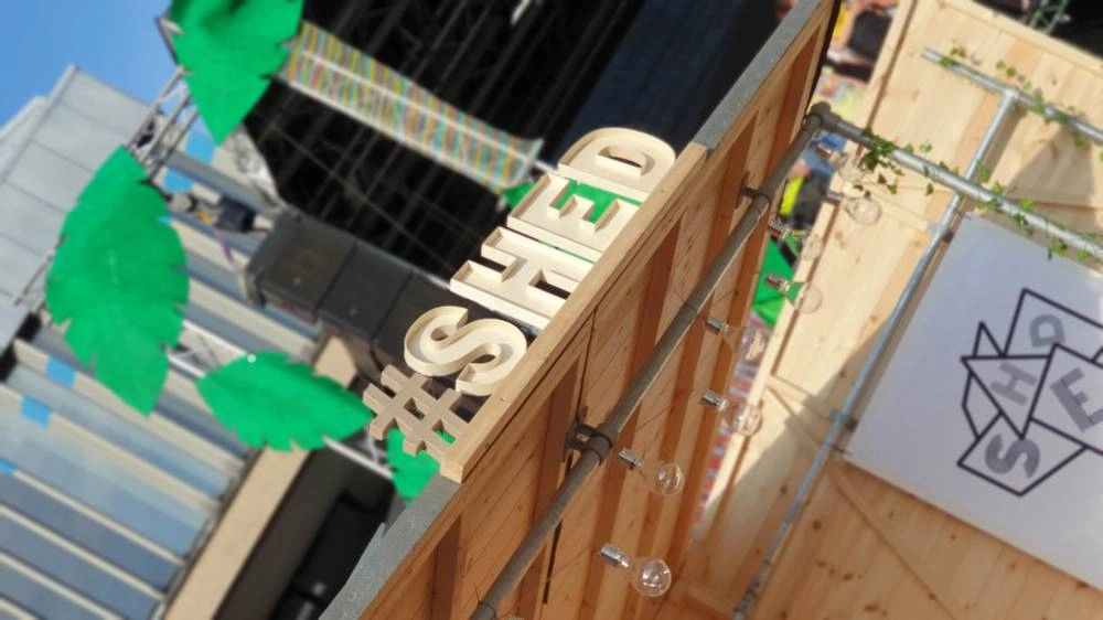Wooden structure with “#SHED” in large letters on top. Green paper leaves decorate the scene. A white sign with a geometric logo is on the right. Urban buildings and a stage are in the background.