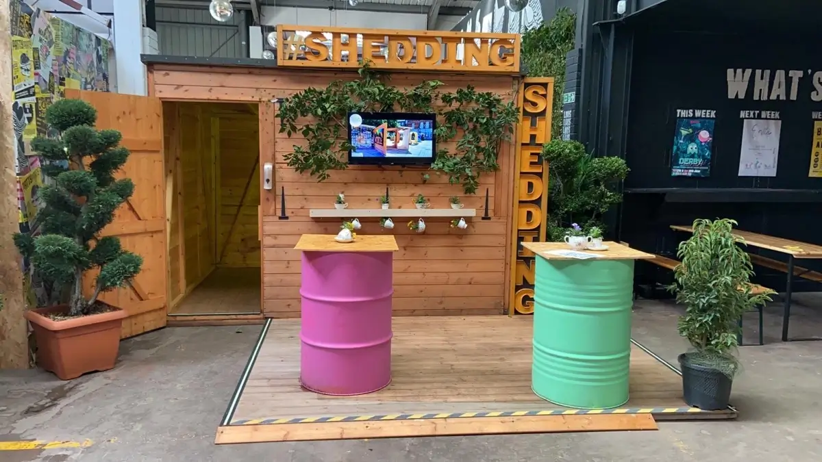 A small wooden shed with SHEDDING signs, greenery, and a TV screen is displayed indoors. In front, there are two tables made from painted barrels—one pink and one green—surrounded by potted plants.