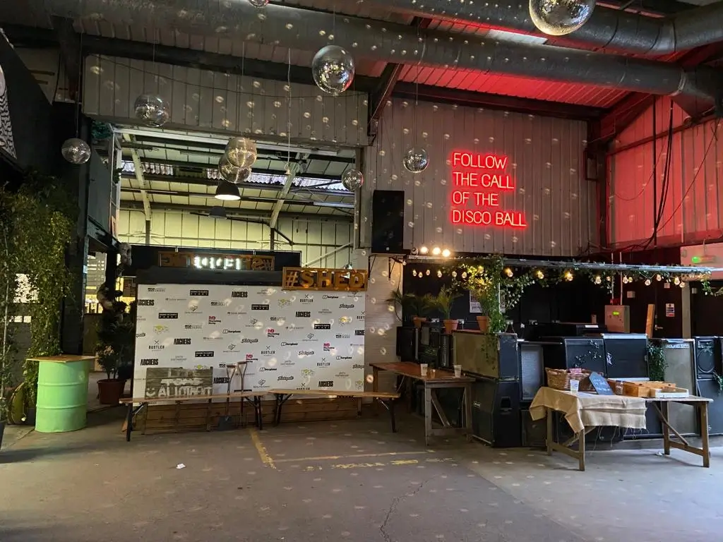 A decorated warehouse event space features a step-and-repeat banner, hashtag sign, tables with food, disco balls, fairy lights, and a neon sign reading FOLLOW THE CALL OF THE DISCO BALL.