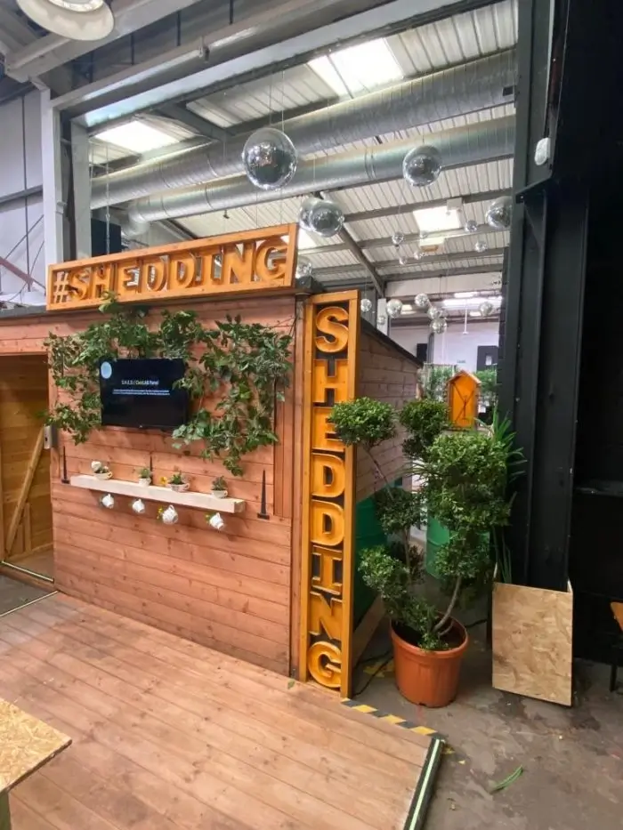 A wooden shed decorated with plants and a TV screen inside a modern indoor space. Large yellow signs read #SHEDDING and SHEDDING. There are disco balls hanging from the ceiling above.