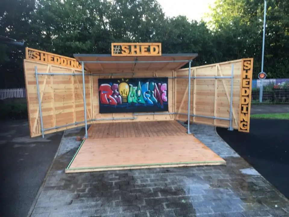 A wooden pop-up shed-like structure with open doors displays colourful graffiti art inside. Signs reading #SHED and #SHEDDING are on the roof and sides. The shed is set up outdoors on a paved surface.