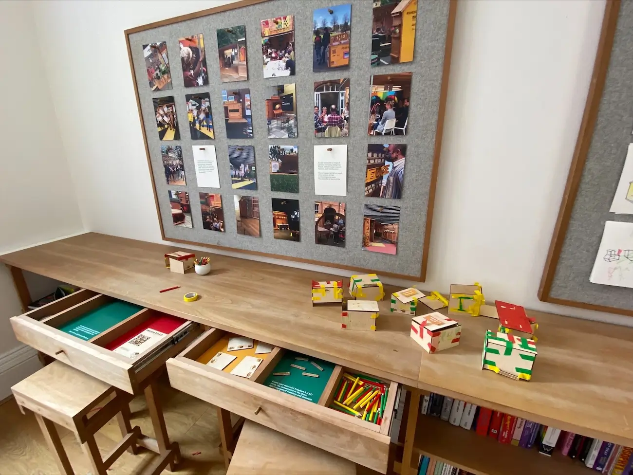 A wooden desk with open drawers containing coloured pencils and paper sits beneath a noticeboard displaying colourful photos and notes. Several small, colourful block constructions are arranged on the desk.