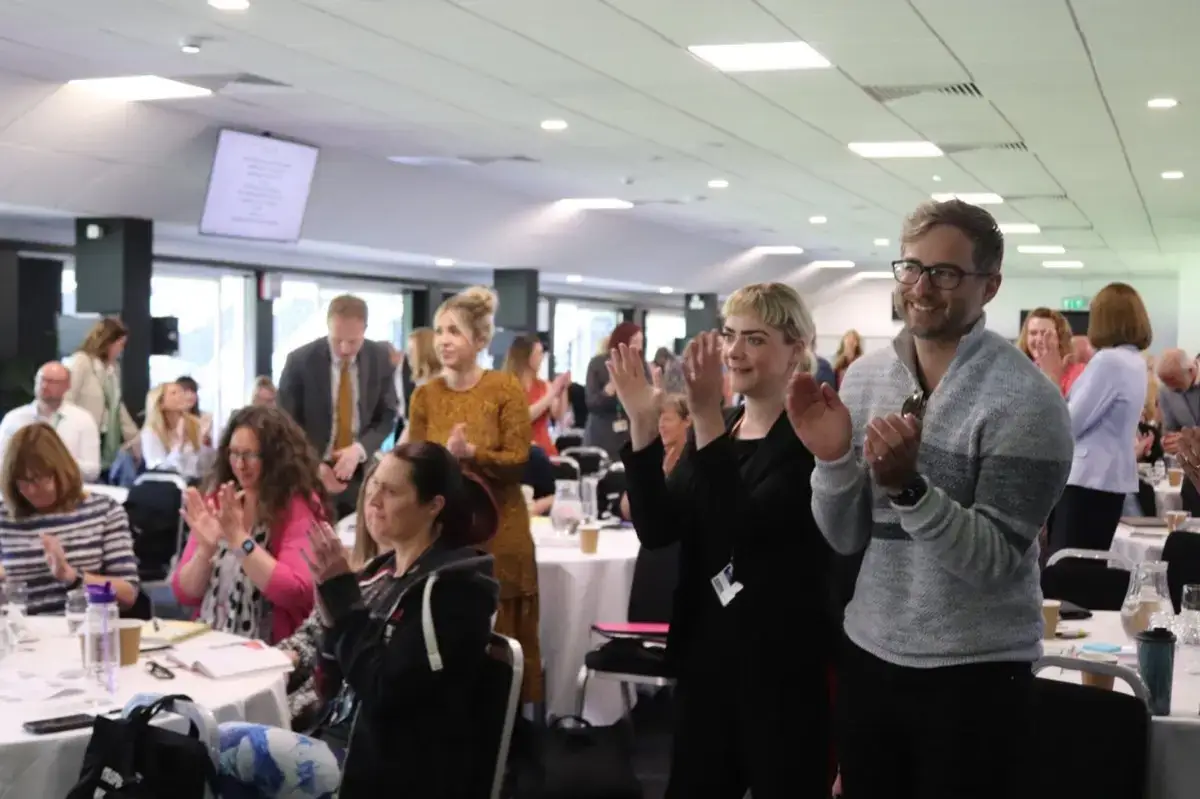 A group of people in a conference room, some seated and some standing, are clapping and smiling during an event. Tables with papers, drinks, and bags are visible in the well-lit room.