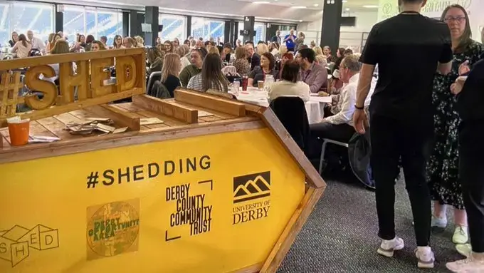A large group of people sit and stand around tables at an indoor event. In the foreground is a yellow sign with hashtags and logos for Shed, Derby County Community Trust, and University of Derby.
