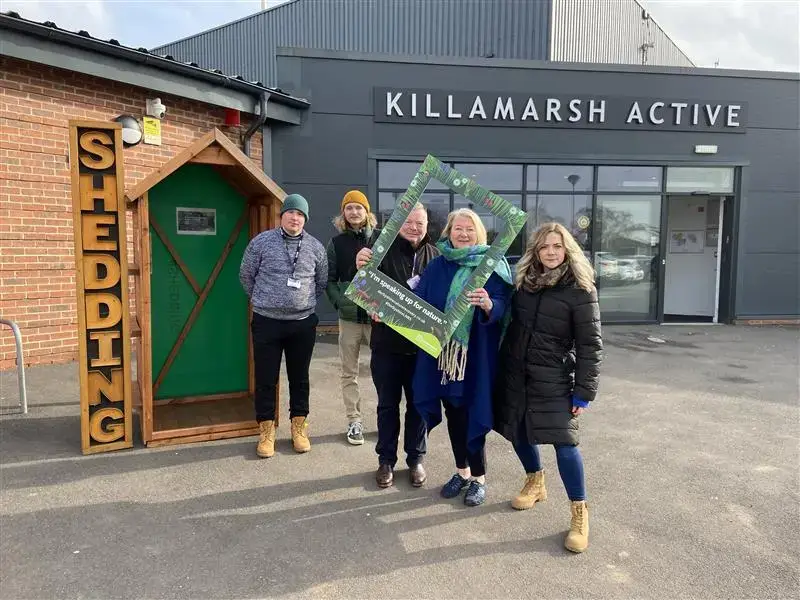 Four people stand outside Killamarsh Active building beside a wooden structure labelled SHEDDING. Two of them hold a large green frame with text, all wearing winter clothing and boots.