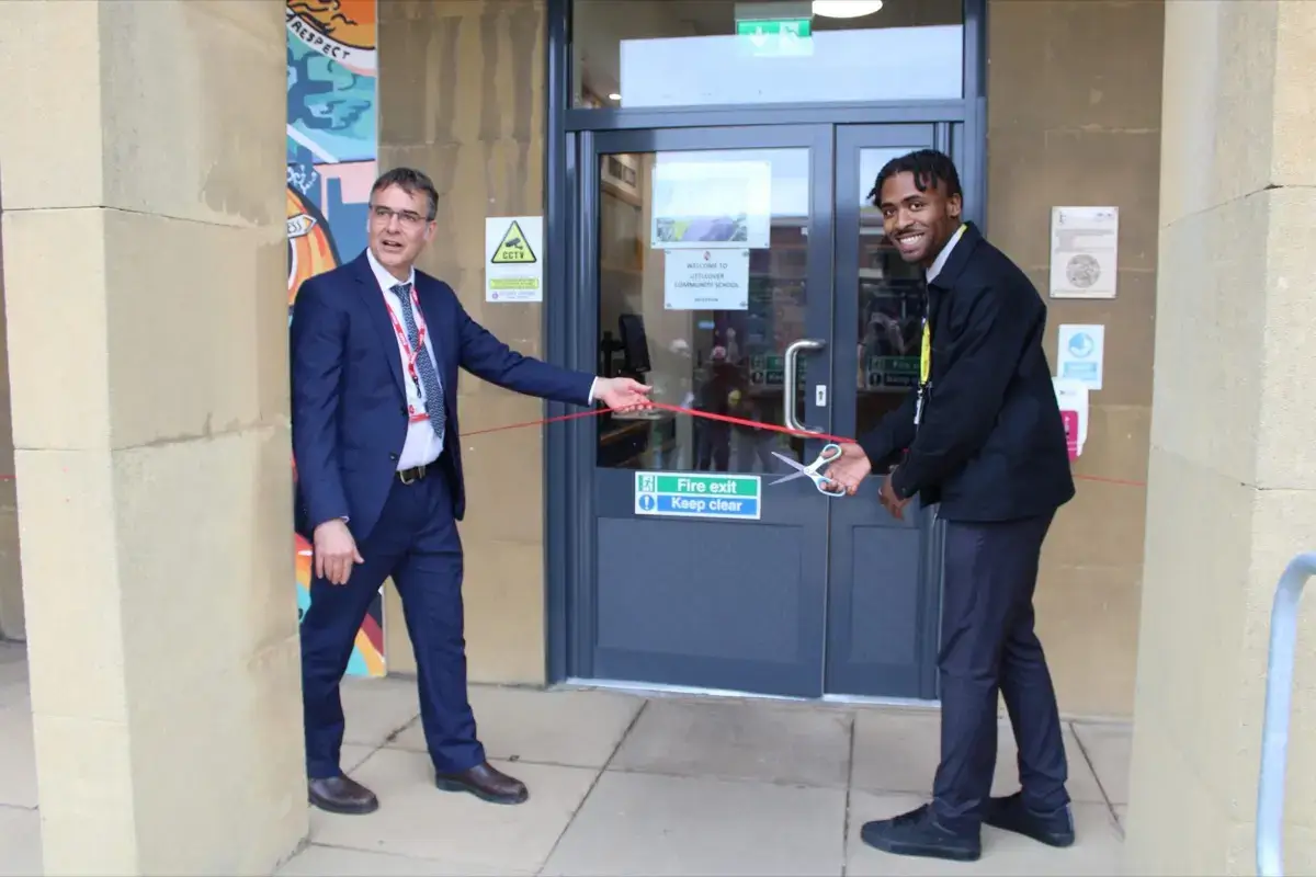 Two men smile as they cut a red ribbon in front of a building entrance, one holding scissors and the other holding the ribbon. The door behind them has a sign that reads Fire exit Keep clear.