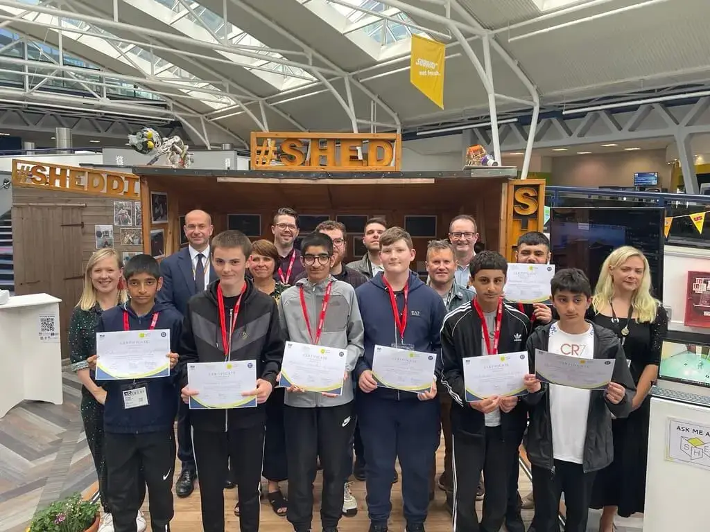 A group of young people stand in front of a wooden stall, each holding certificates. Adults stand behind them, smiling. The setting appears to be indoors, possibly at an event or awards ceremony.