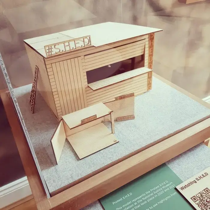 A wooden architectural model of a small modern building labelled #SHED, featuring large rectangular windows, a ramp, and moveable panels, displayed on a flat surface with a green information plaque nearby.