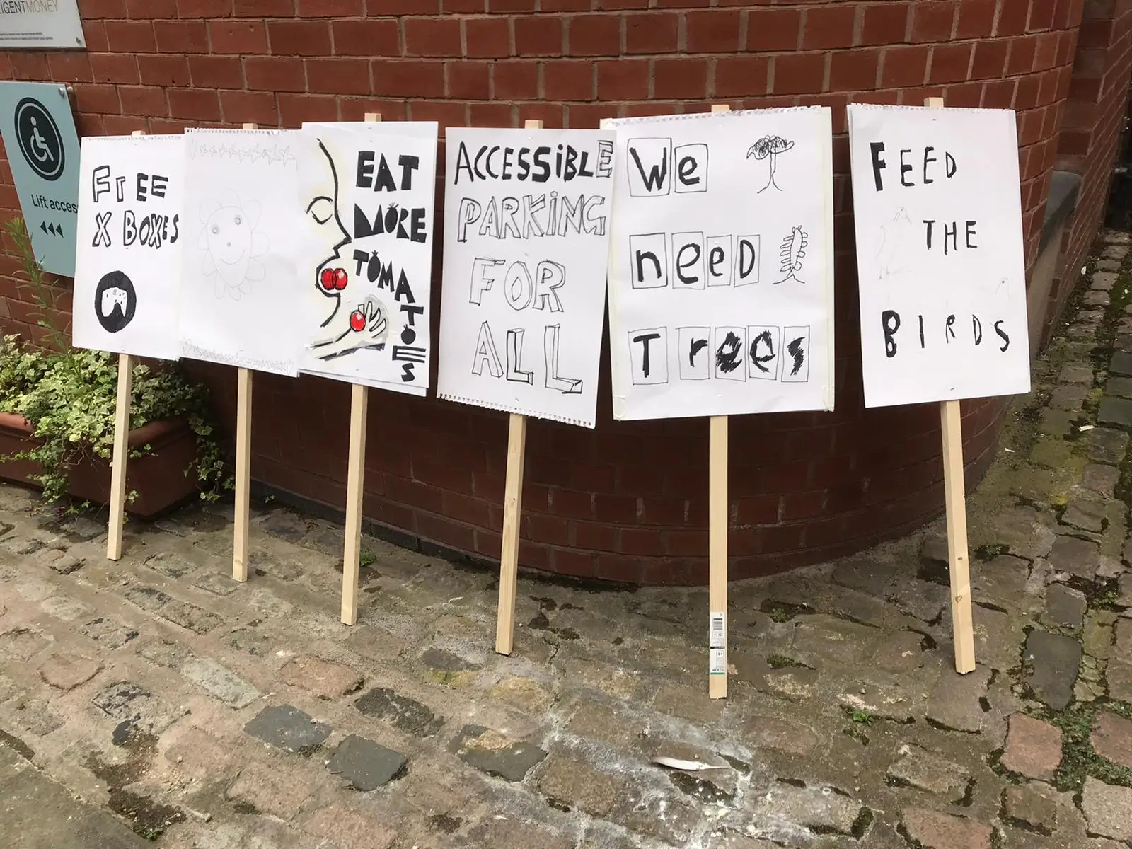 Six hand-drawn protest signs on sticks lean against a brick wall, displaying messages such as “Free x Books”, “Eat More Tuna”, “Accessible Parking for All”, “We Need Trees” and “Feed the Birds”.
