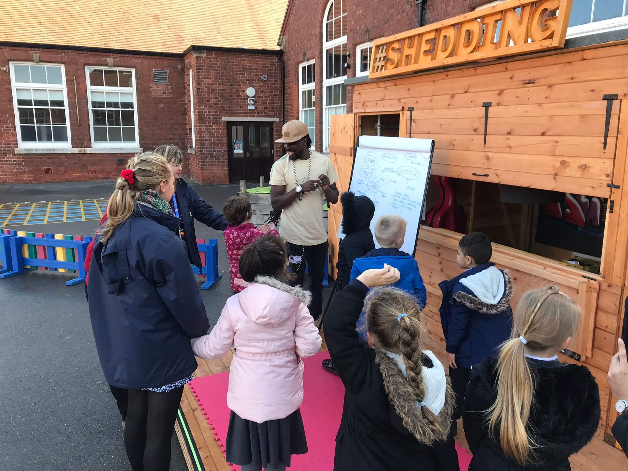 A man stands by a whiteboard outside a wooden shed, talking to a group of children and two adults. The children, wearing coats, listen attentively in a schoolyard. The shed has a #SHEDDING sign.