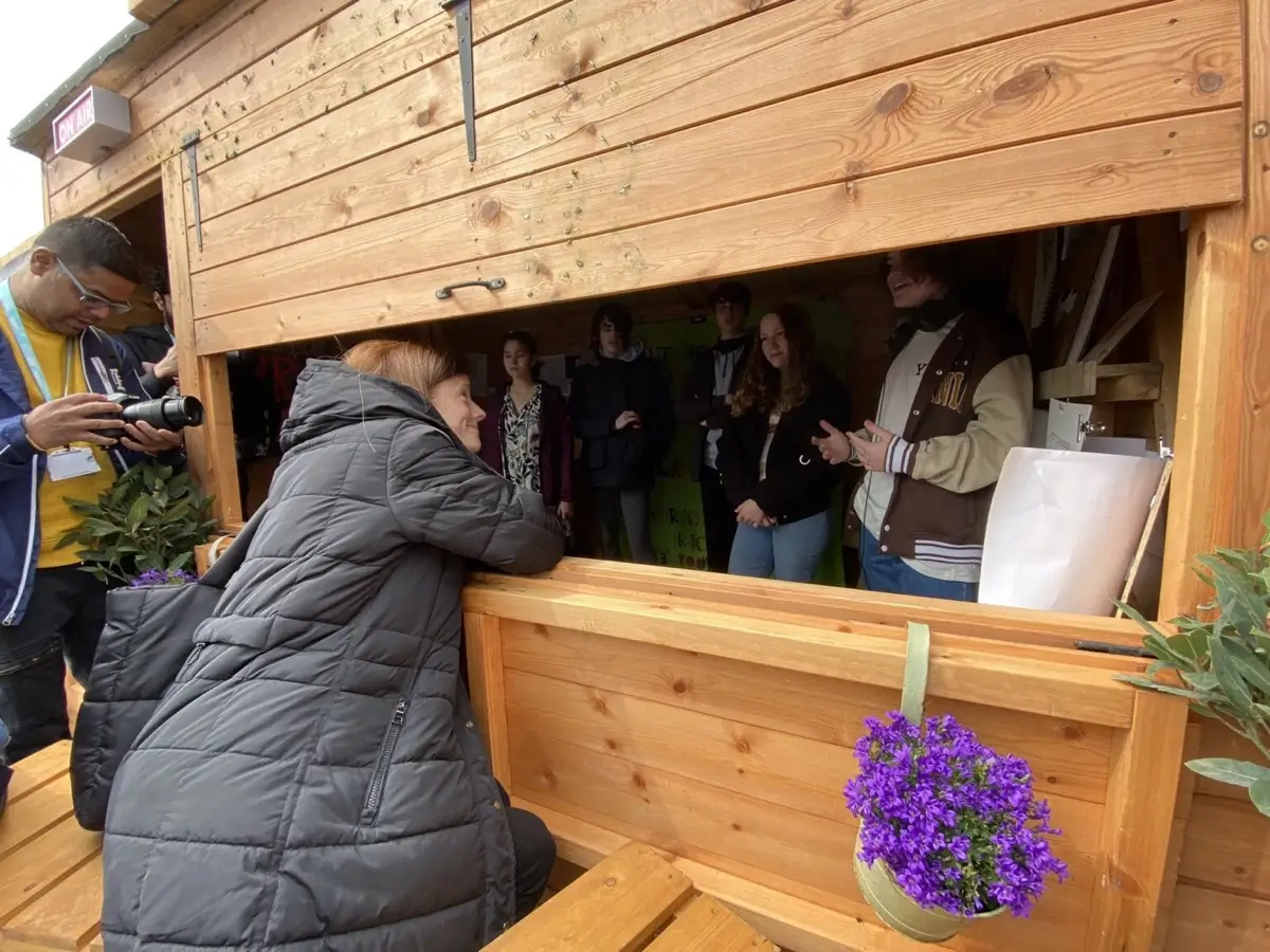 A woman in a black coat leans on a wooden counter, listening to a group of people inside a wooden booth. A man with a camera stands nearby. Purple flowers decorate the counter.