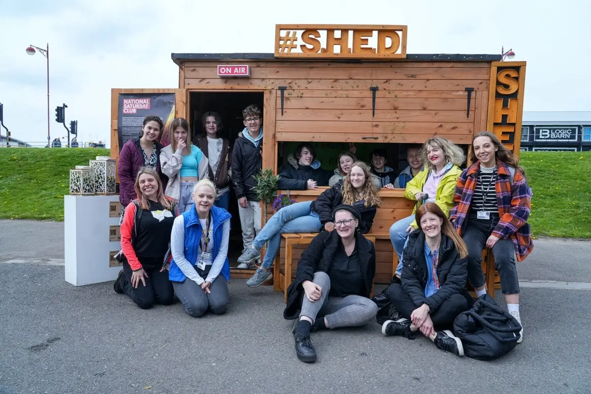 A group of people pose and smile in front of and inside a wooden shed labelled "#SHED" on a paved area outdoors.