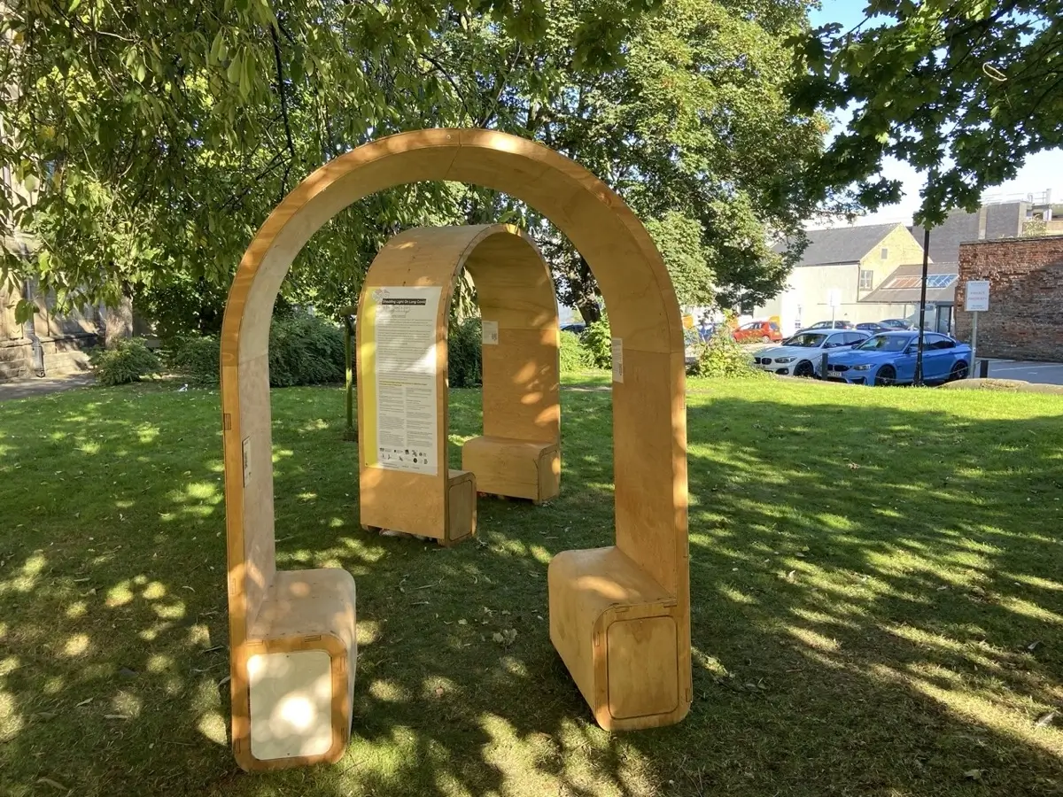 A wooden arched structure with benches underneath stands on grass in a shaded park area, with informational signs attached and several parked cars and buildings visible in the background.
