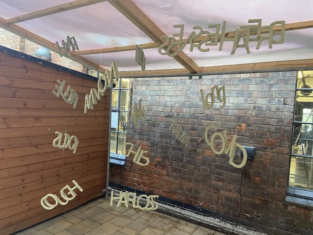 Gold letters hang from the ceiling inside a wooden and brick structure, spelling words that are reversed and appear to float in mid-air. The words are reflected in the glass window on the right.
