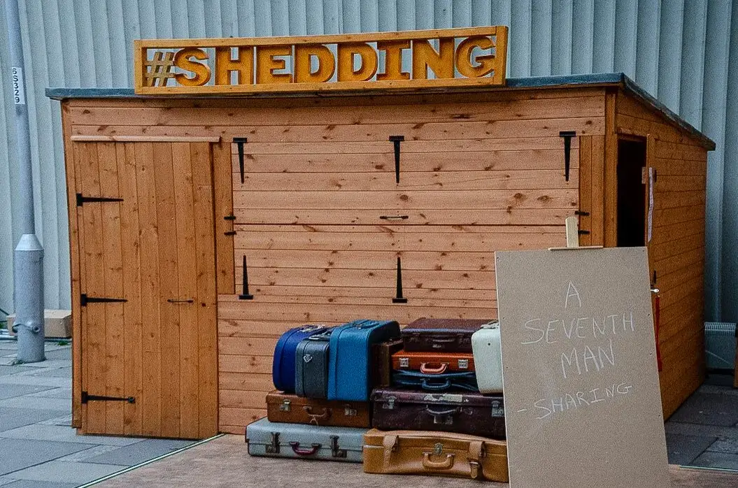 A wooden shed with a sign reading #SHEDDING on the roof. Several vintage suitcases are stacked outside, next to a cardboard sign that says A SEVENTH MAN - SHARING. The scene is outdoors.