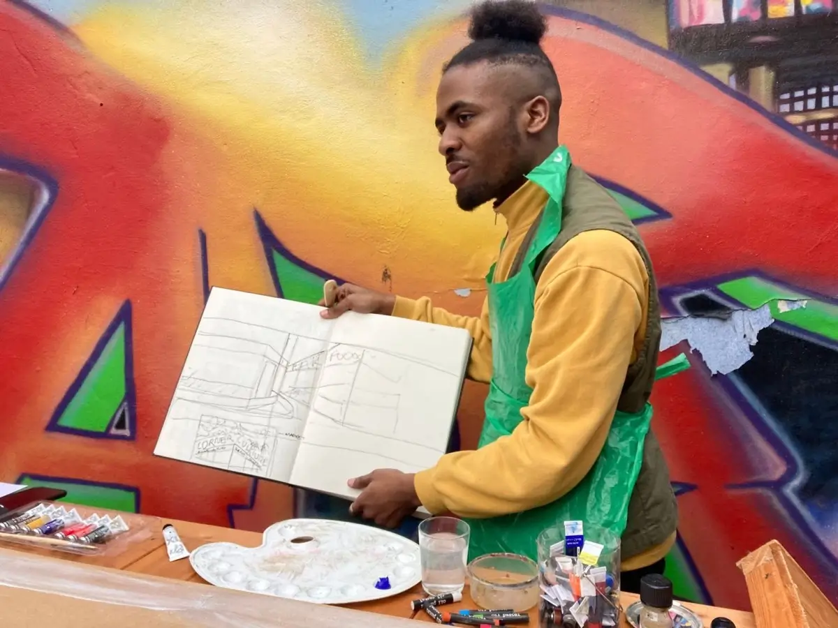 A man in a green apron holds up a sketchbook displaying a pencil drawing. He stands in front of a colourful graffiti wall, with art supplies like paint tubes and brushes scattered on the table in front of him.