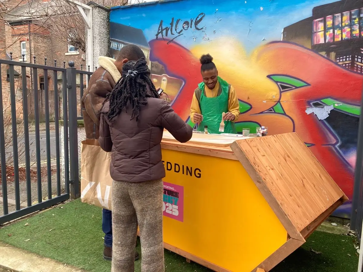 Two people stand at a yellow outdoor wooden stall, speaking to a vendor in a green apron. The background features a colourful graffiti mural and buildings. The scene appears to be in an urban area.