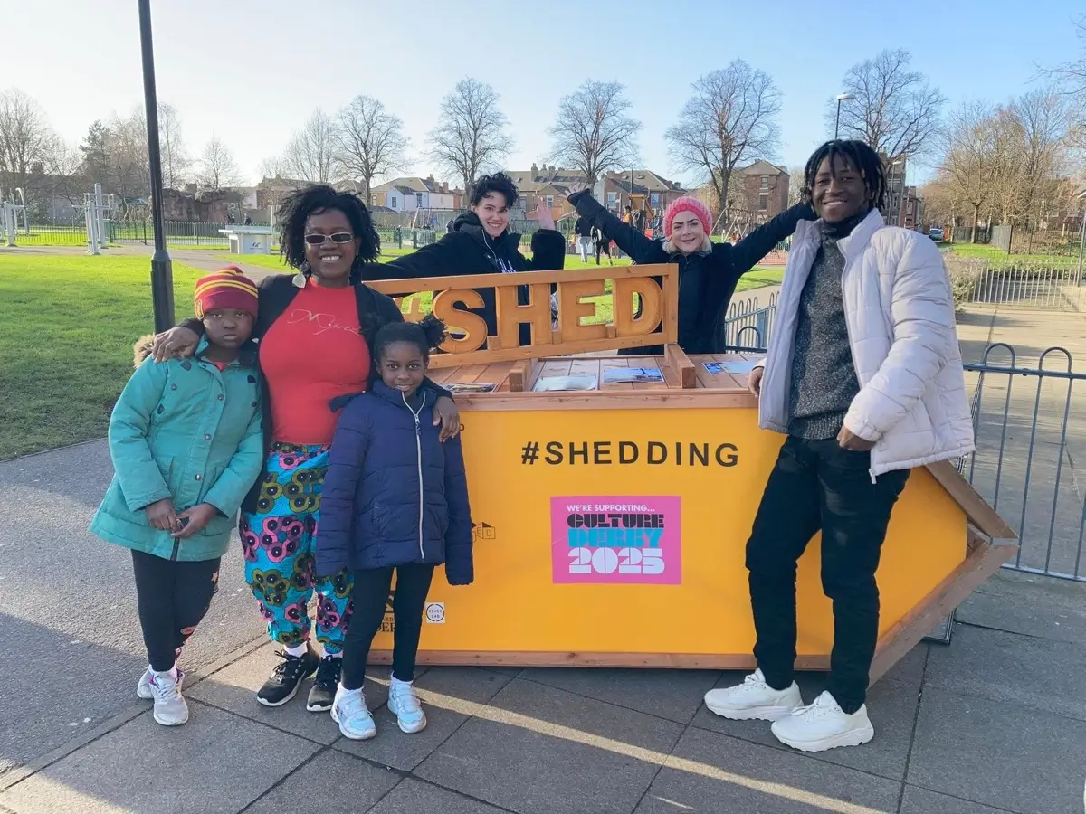 A group of six people, including children and adults, smile and pose in front of a large yellow structure labelled SHED in a park. A pink sign reads “#SHEDDING – Culture Derby 2025.” The weather is sunny and clear.