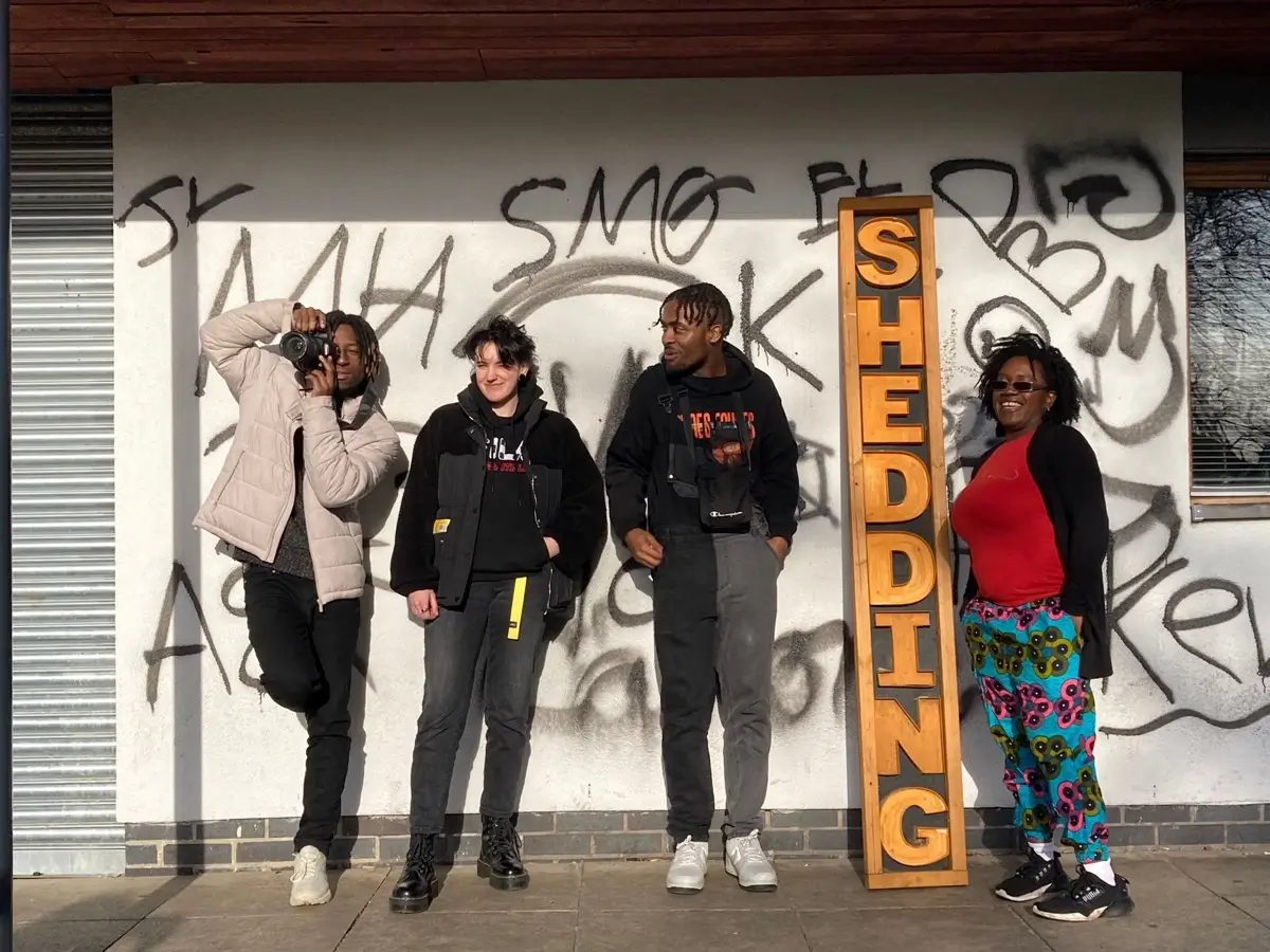 Four people stand in front of a graffiti-covered wall, posing casually next to a tall wooden sign that reads SHEDDING in bold letters. One person is taking a photo, and all appear relaxed and happy.