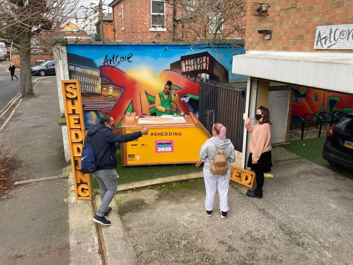Three people stand outside near a yellow kiosk labelled SHEDDING, speaking to a person behind the counter. Colourful street art decorates the wall behind them, and large vertical text reads SHEDDING.