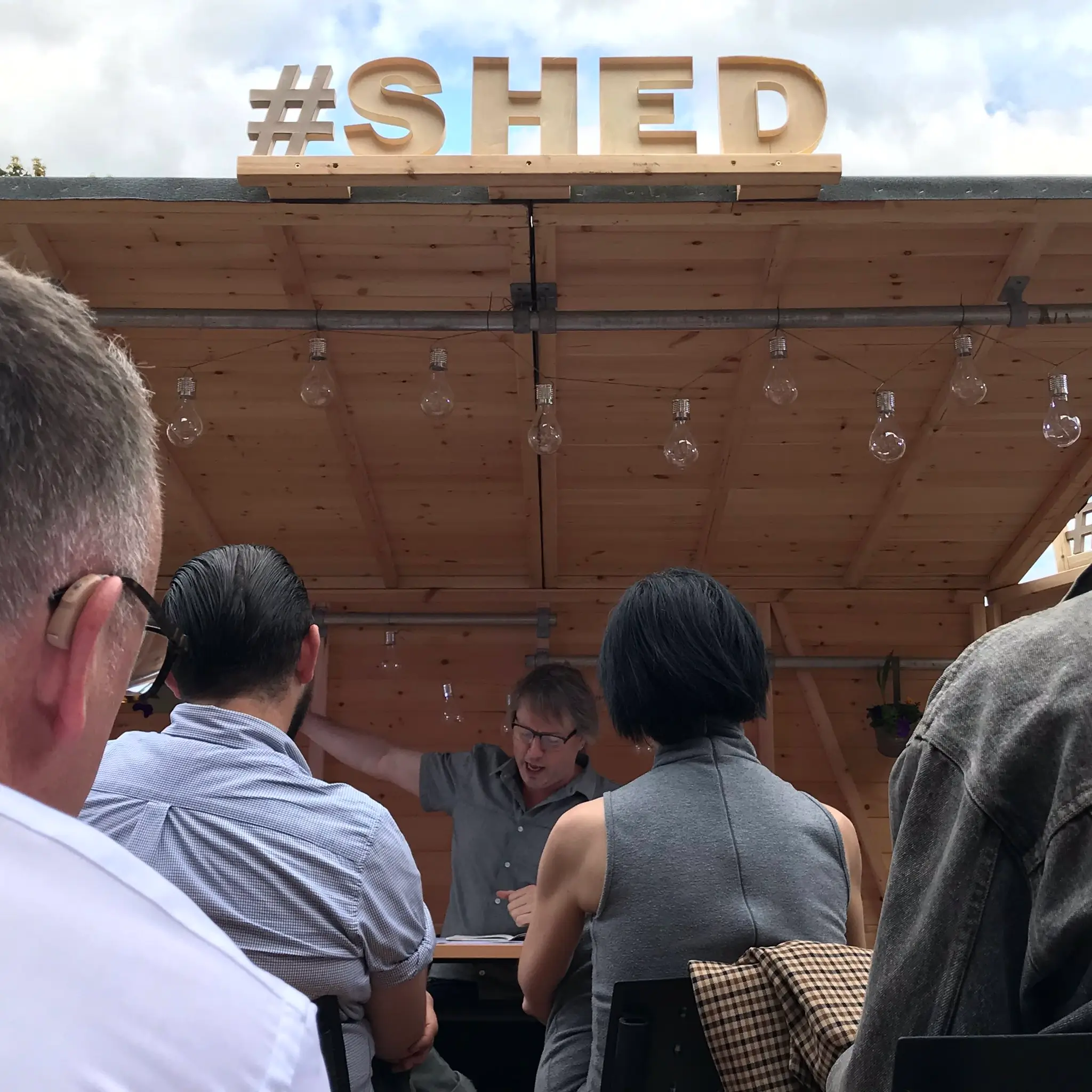 A person speaks enthusiastically at an outdoor event under a wooden shelter with a large #SHED sign on top, facing an audience seated with their backs to the camera. Light bulbs hang from the shelter roof.