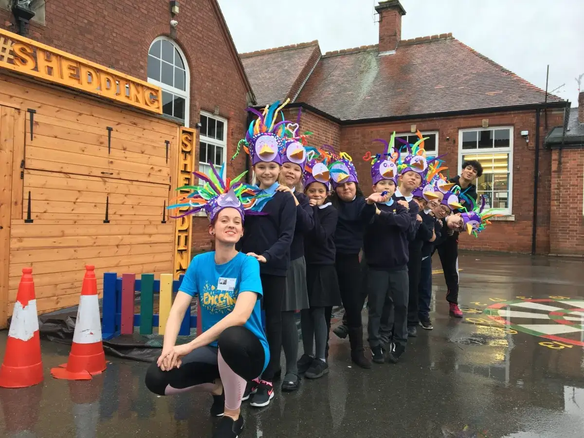 A group of children wearing colourful, handmade unicorn hats stand in a queue outside a brick building, smiling. An adult in a blue shirt kneels in front, and the ground is wet, reflecting the building and cones nearby.
