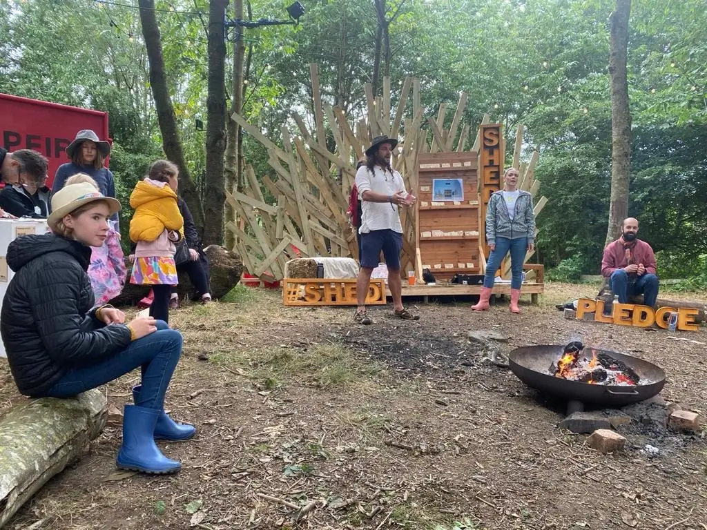 A group of people sit and stand around a campfire in a wooded area. Some wear wellington boots and hats. A person in the centre speaks, holding papers, near a wooden backdrop with signs reading “SHED” and “PLEDGE”.