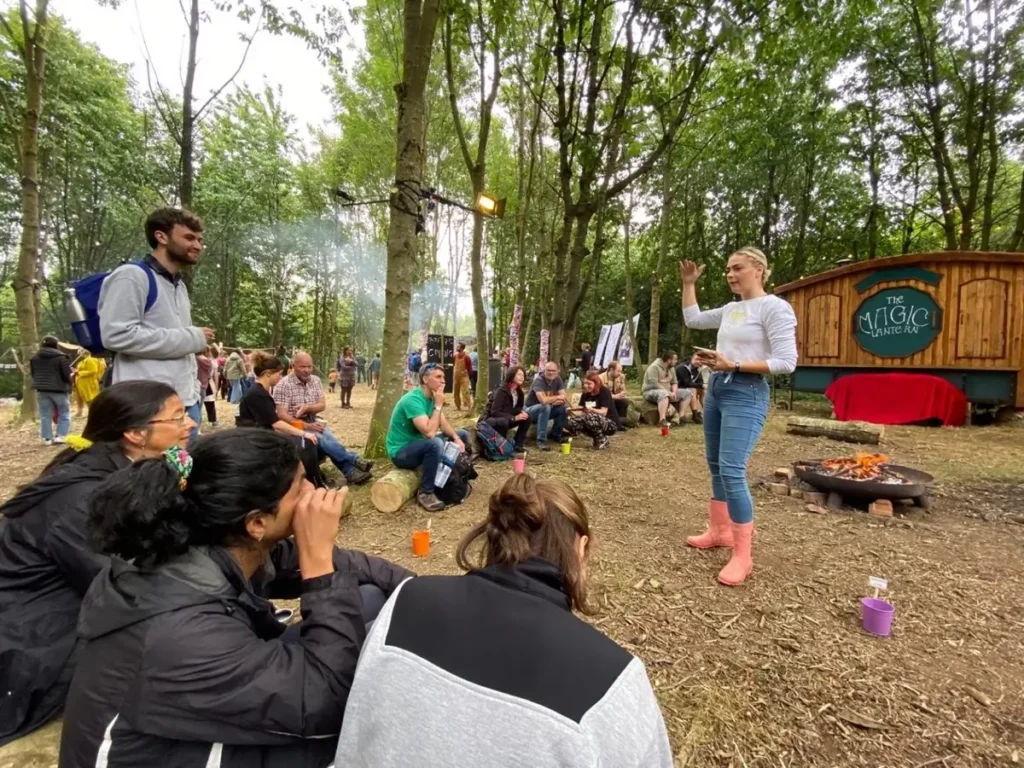 A woman stands and speaks to a seated group outdoors in a wooded area near a firepit. People listen attentively. Behind them is a wooden hut labelled The Magic Campervan. Colourful cups are on the ground.