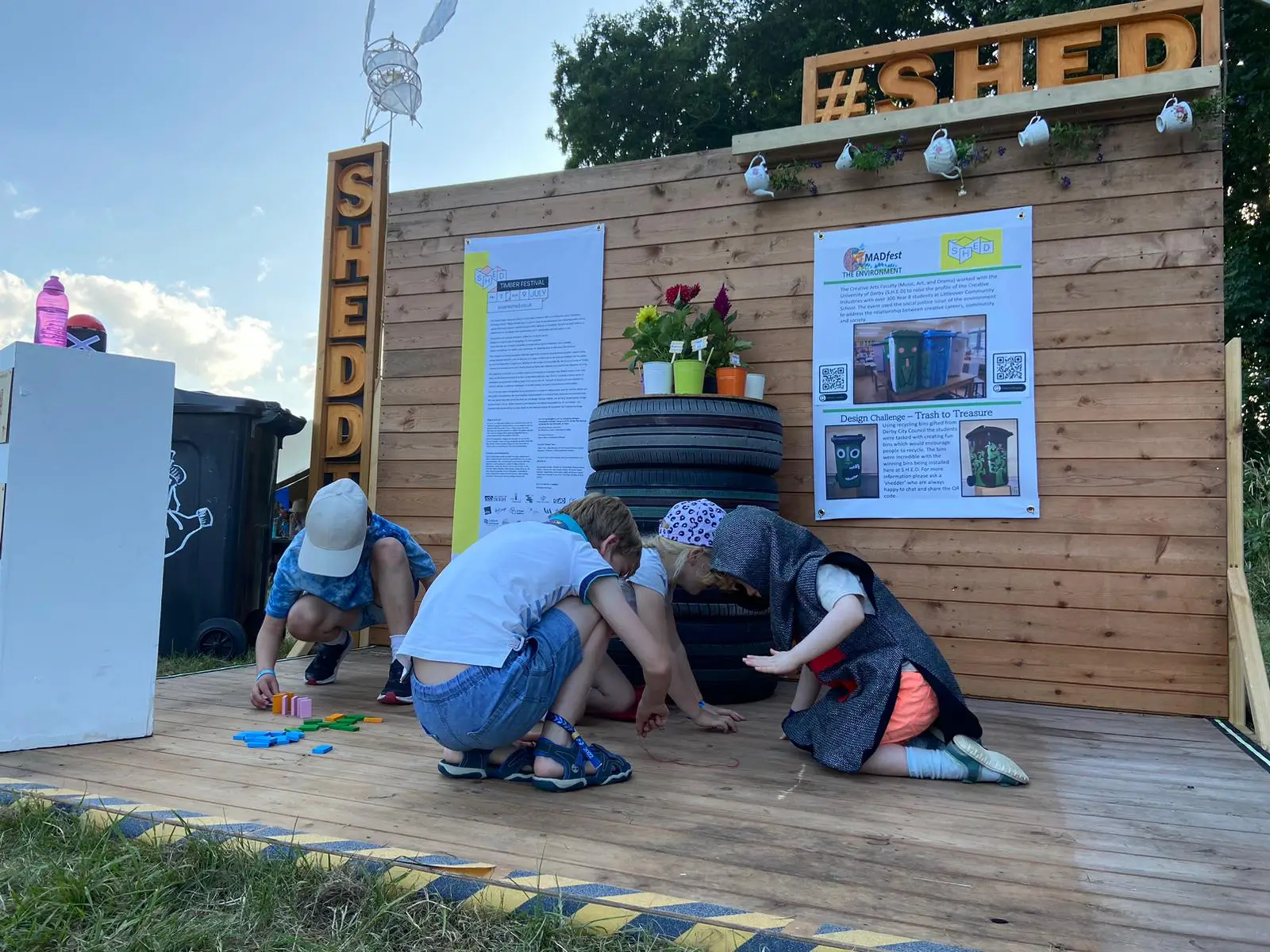 Three children play and explore on a wooden platform in front of a wood-panelled outdoor display with signage, potted plants, and recycled tyre planters at a community or environmental event.