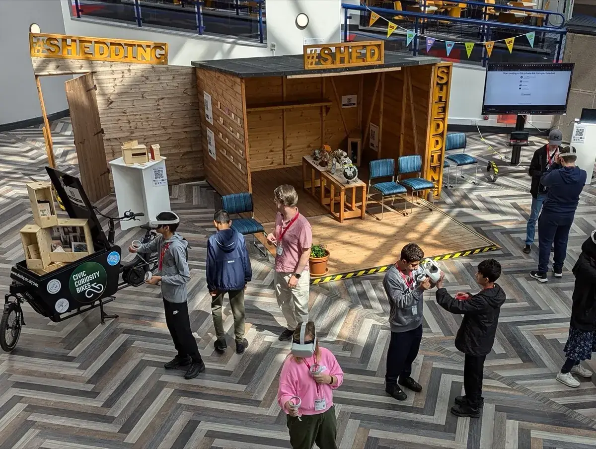 A group of people, some wearing VR headsets, explore an exhibit called Shedding Shed in a large indoor space with patterned flooring. The exhibit features a wooden shed, decorations, and interactive displays.