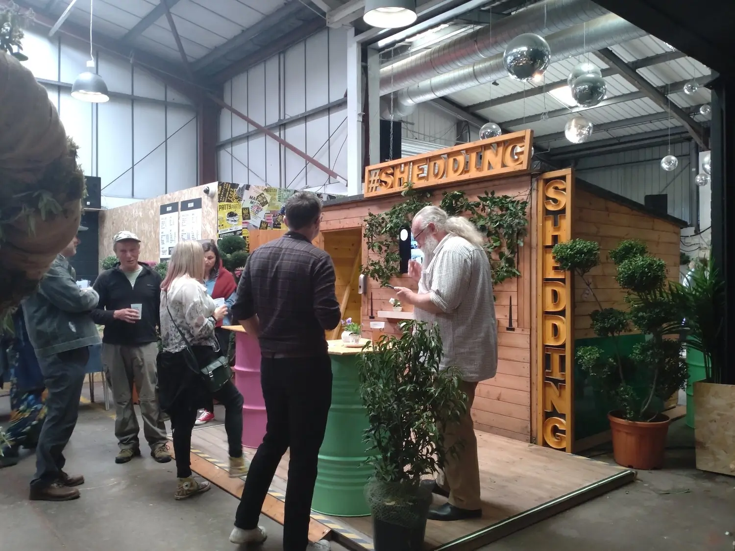 People gather and talk in front of a wooden stall labelled SHEDDING at an indoor event. The stall is decorated with plants, and barrels serve as tables. The setting is industrial with exposed beams and ducts.